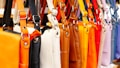 A row of colorful leather handbags displayed in a variety of vibrant shades ranging from orange, black, white, and brown to yellow, red, purple, and pink. The bags are neatly hung by their straps, highlighting their design and metallic buckles.