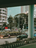 A bustling urban street scene featuring a multi-story building, a large sign reading 'Kandivli', and various vehicles, including a motorcycle and cars waiting at a traffic signal. The area is lined with trees and greenery, with people walking on the sidewalk.