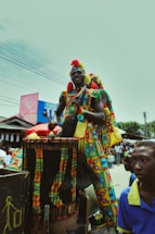 A person dressed in colorful traditional attire stands on a platform, playing a large drum. Surrounding them are other individuals in vibrant clothing, with buildings and crowd visible in the background.