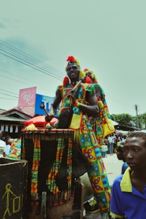 A person dressed in colorful traditional attire stands on a platform, playing a large drum. Surrounding them are other individuals in vibrant clothing, with buildings and crowd visible in the background.