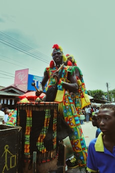 A person dressed in colorful traditional attire stands on a platform, playing a large drum. Surrounding them are other individuals in vibrant clothing, with buildings and crowd visible in the background.