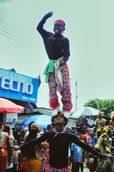 Two performers are balancing skillfully in a lively street setting, with one cheerfully standing on top of the other’s head. They are wearing bright, colorful costumes adorned with patterns. The background is filled with people observing, along with vibrant banners and signage, adding to the festive atmosphere.