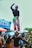 Two performers are balancing skillfully in a lively street setting, with one cheerfully standing on top of the other’s head. They are wearing bright, colorful costumes adorned with patterns. The background is filled with people observing, along with vibrant banners and signage, adding to the festive atmosphere.