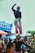 Two performers are balancing skillfully in a lively street setting, with one cheerfully standing on top of the other’s head. They are wearing bright, colorful costumes adorned with patterns. The background is filled with people observing, along with vibrant banners and signage, adding to the festive atmosphere.