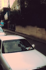 A white car is parked on a street lined with residential buildings. The windshield has a sticker with some text and numbers, and trees cast long shadows on the car. The buildings in the background are partially obscured by trees and bushes.