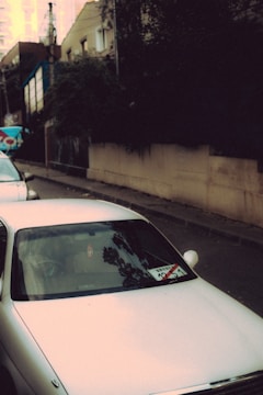 A white car is parked on a street lined with residential buildings. The windshield has a sticker with some text and numbers, and trees cast long shadows on the car. The buildings in the background are partially obscured by trees and bushes.