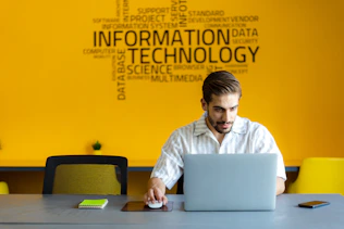 A man is seated at a table, working on a laptop in front of a bright yellow wall. The wall has a large collection of words related to technology, such as 'Information Technology' and 'Data'. A chair with a striped design is partially visible, and a notebook and phone rest on the table.