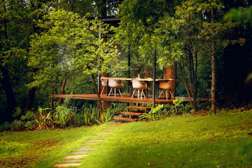 A serene setting featuring a small, elevated wooden deck surrounded by lush green trees. The deck contains a table with several stylish chairs, likely designed for outdoor dining or relaxation. A stone path leads up to the deck, enhancing the natural and tranquil atmosphere.