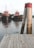 Two red tugboats float side by side in a calm body of water, with a wooden dock prominently in the foreground. A pair of weathered wooden posts with red tops stands near the edge of the dock. The background features greenery and a cloudy sky, contributing to a tranquil setting.