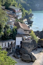 A coastal scene with several houses built on a rocky shoreline. The buildings feature a mix of modern and traditional architecture, some with thatched roofs and balconies overlooking the sea. Lush greenery surrounds the area, and the turquoise water gently laps against the rocks. A small sandy beach at the bottom is partially covered by shadows from the adjacent cliffs.