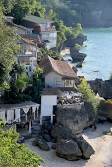 A coastal scene with several houses built on a rocky shoreline. The buildings feature a mix of modern and traditional architecture, some with thatched roofs and balconies overlooking the sea. Lush greenery surrounds the area, and the turquoise water gently laps against the rocks. A small sandy beach at the bottom is partially covered by shadows from the adjacent cliffs.