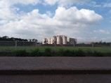 A large, modern architectural structure sits behind a wide expanse of grass and a fence. The sky is filled with white, fluffy clouds under a blue background. Trees are visible in the distance, framing the building.