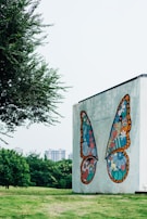 A large wall mural depicts vibrantly colored, symmetrical butterfly wings composed of intricate designs and shapes, including hearts, flowers, and a small rainbow. The mural is set against a pale concrete wall. Nearby, lush green grass and trees add natural elements to the urban setting, while distant high-rise buildings are visible through a light haze.