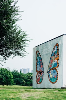 A large wall mural depicts vibrantly colored, symmetrical butterfly wings composed of intricate designs and shapes, including hearts, flowers, and a small rainbow. The mural is set against a pale concrete wall. Nearby, lush green grass and trees add natural elements to the urban setting, while distant high-rise buildings are visible through a light haze.