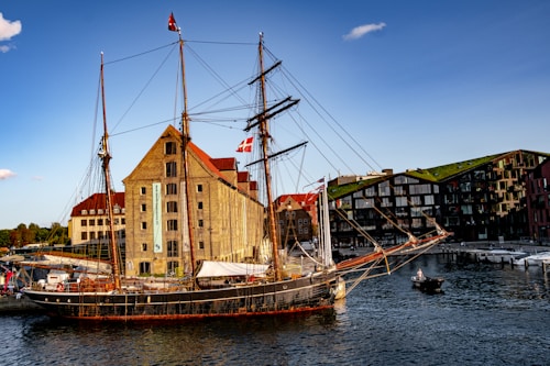 A tall ship is docked alongside a historic brick building with a red roof. The ship has multiple masts with intricate rigging and flies a flag. Nearby, there is a modern building with a green rooftop. A small boat with two people is in the water.
