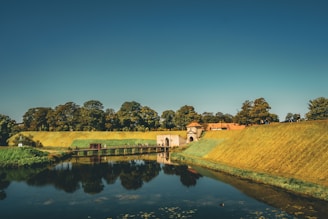 A serene landscape featuring a historic building with a red-tiled roof sitting atop a gently sloped grassy hill. The scene includes a reflective body of water in the foreground, bordered by lush greenery and tall trees lining the horizon. The sky above is clear with a deep blue hue.