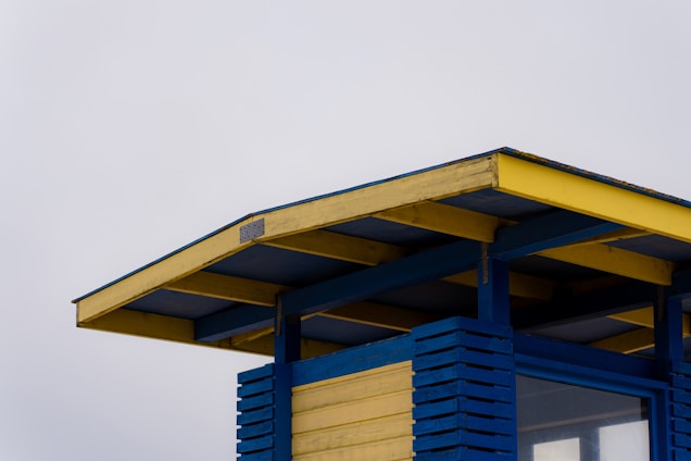 A structure with a roof made of wooden beams painted in blue and yellow, featuring an overhang. The walls of the structure have a slatted design with blue and yellow planks. The sky in the background is overcast and light gray.
