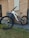 A mountain bike with a sturdy frame is positioned against a brick wall next to a large gray bin. The bicycle features thick, durable tires branded with 'Maxxis' and is equipped with a suspension fork. The setting includes elements of a paved area and a section of grass in the foreground.