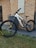A mountain bike with a sturdy frame is positioned against a brick wall next to a large gray bin. The bicycle features thick, durable tires branded with 'Maxxis' and is equipped with a suspension fork. The setting includes elements of a paved area and a section of grass in the foreground.