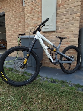 A mountain bike with a sturdy frame is positioned against a brick wall next to a large gray bin. The bicycle features thick, durable tires branded with 'Maxxis' and is equipped with a suspension fork. The setting includes elements of a paved area and a section of grass in the foreground.
