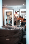 A person is getting their hair styled in a modern salon. The stylist, wearing a striped dress, is focusing on the client's hair while using a comb. Mirrors in the background reflect the interior, containing shelves with various hair products and a decorative plant. The overall setting is clean and well-lit, with multiple light sources providing a bright and inviting atmosphere.