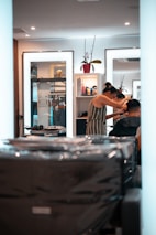 A person is getting their hair styled in a modern salon. The stylist, wearing a striped dress, is focusing on the client's hair while using a comb. Mirrors in the background reflect the interior, containing shelves with various hair products and a decorative plant. The overall setting is clean and well-lit, with multiple light sources providing a bright and inviting atmosphere.