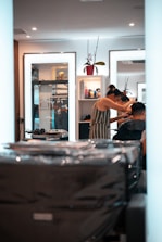 A person is getting their hair styled in a modern salon. The stylist, wearing a striped dress, is focusing on the client's hair while using a comb. Mirrors in the background reflect the interior, containing shelves with various hair products and a decorative plant. The overall setting is clean and well-lit, with multiple light sources providing a bright and inviting atmosphere.