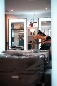 A person is getting their hair styled in a modern salon. The stylist, wearing a striped dress, is focusing on the client's hair while using a comb. Mirrors in the background reflect the interior, containing shelves with various hair products and a decorative plant. The overall setting is clean and well-lit, with multiple light sources providing a bright and inviting atmosphere.