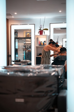 A person is getting their hair styled in a modern salon. The stylist, wearing a striped dress, is focusing on the client's hair while using a comb. Mirrors in the background reflect the interior, containing shelves with various hair products and a decorative plant. The overall setting is clean and well-lit, with multiple light sources providing a bright and inviting atmosphere.