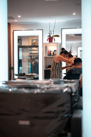 A person is getting their hair styled in a modern salon. The stylist, wearing a striped dress, is focusing on the client's hair while using a comb. Mirrors in the background reflect the interior, containing shelves with various hair products and a decorative plant. The overall setting is clean and well-lit, with multiple light sources providing a bright and inviting atmosphere.