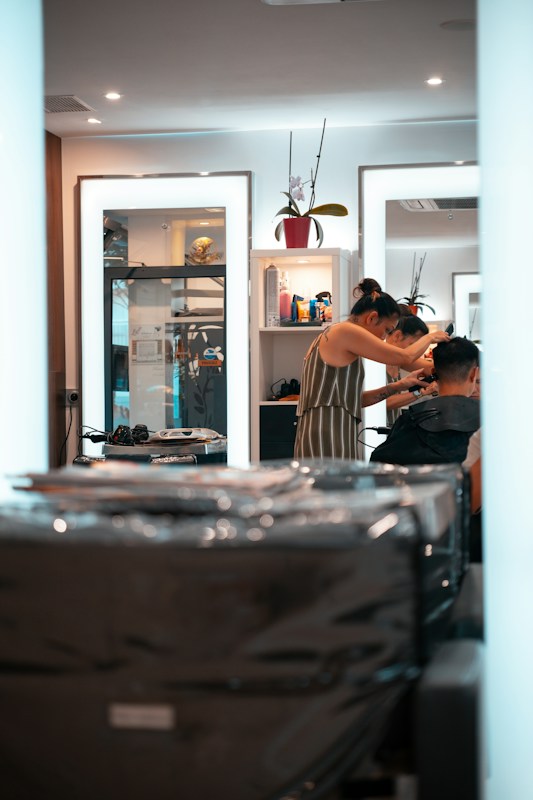 A person is getting their hair styled in a modern salon. The stylist, wearing a striped dress, is focusing on the client's hair while using a comb. Mirrors in the background reflect the interior, containing shelves with various hair products and a decorative plant. The overall setting is clean and well-lit, with multiple light sources providing a bright and inviting atmosphere.
