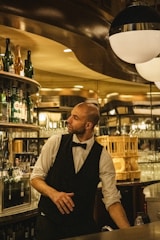 A bartender wearing a black vest and white shirt with a bow tie stands behind a bar counter, surrounded by various bottles and glassware. He appears to be in a restaurant or bar setting, with warm lighting creating a cozy ambiance.