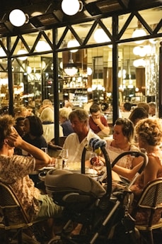A bustling indoor cafe setting filled with people engaged in conversation and dining. The ambiance is warm and inviting, with several globe lights hanging from the ceiling and illuminating the space. Patrons are seated closely at tables, reflecting a lively and social atmosphere. A stroller is parked next to a table where people are enjoying their meals.