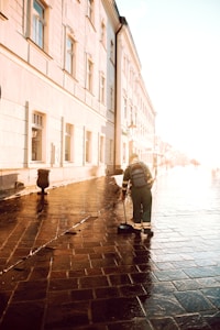 A person wearing a uniform is sweeping a wet cobblestone street next to a row of historic buildings. The sunlight creates a warm glow on the buildings, enhancing the texture of the bricks. A trash can and a street lamp are visible along the sidewalk.