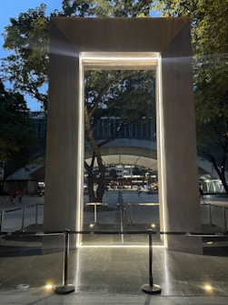 An illuminated architectural frame stands in an outdoor urban setting, bordered by trees. The frame is large and metallic, with bright lighting outlining its rectangular shape. In the background, a large canopy structure and people walking across a pathway add to the city's bustling atmosphere.