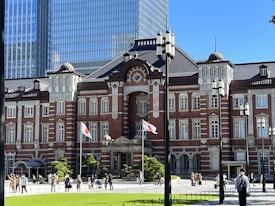 A historic red brick building with ornate architectural details, featuring multiple windows and decorative columns, is set against a backdrop of modern glass skyscrapers. A neatly manicured garden with small trees and a green lawn is in the foreground, where people are walking casually. Two flagpoles displaying Japanese flags are visible near the entrance of the building.