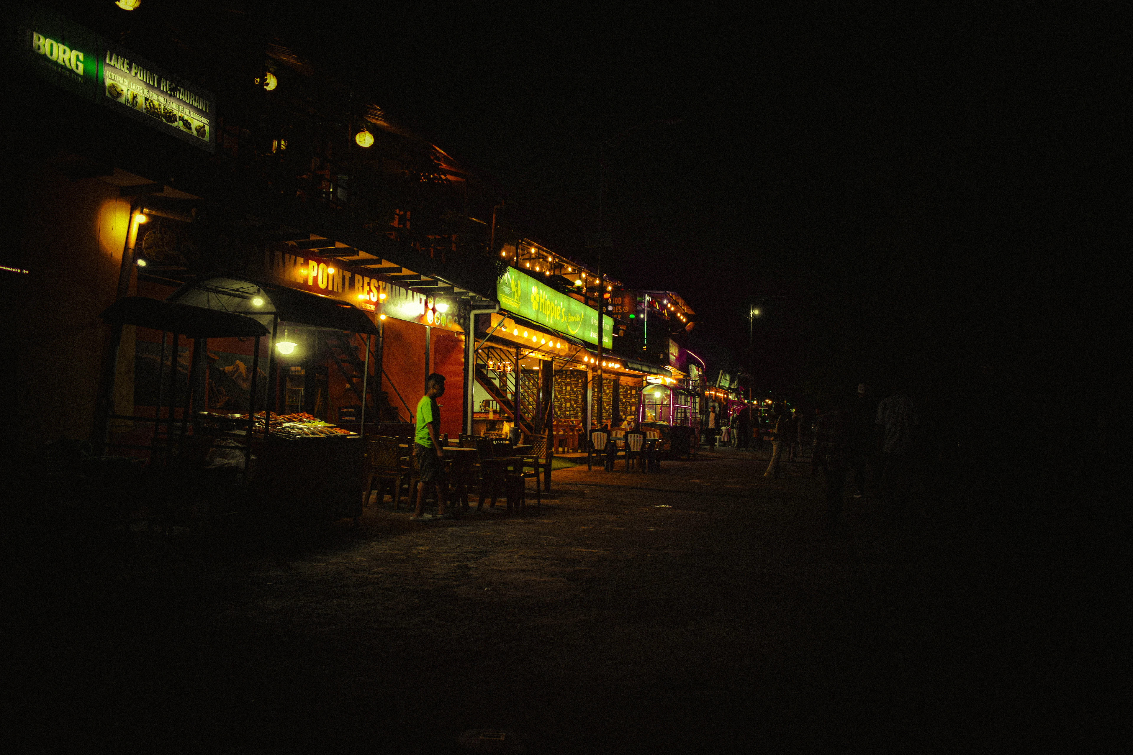 The Lakeside strip in Pokhara after dark, a row of open-fronted restaurants glowing with string lights and green signboards along an unlit road, one figure crossing between the tables