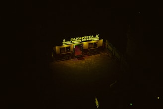 A dimly lit restaurant with a yellow sign reading 'Janapriya Thakali Bhancha Ghar & Restaurant' is visible. The sign is illuminated against a dark background, and there are some plants and posters near the entrance.