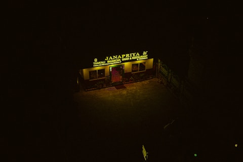 A dimly lit restaurant with a yellow sign reading 'Janapriya Thakali Bhancha Ghar & Restaurant' is visible. The sign is illuminated against a dark background, and there are some plants and posters near the entrance.