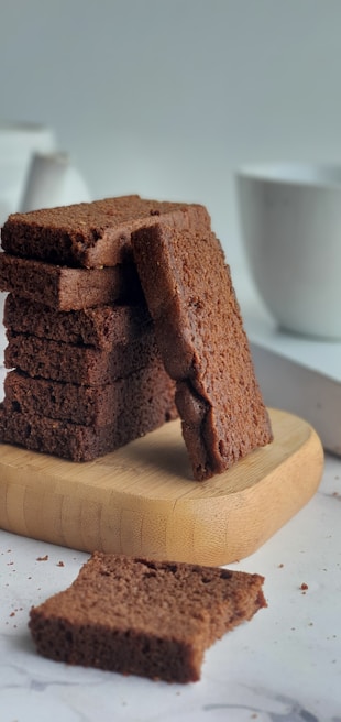A stack of golden pound cake slices on a rustic wooden board with pastel pink and purple baking tools in the background