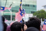 A crowd of people holding and waving Malaysian flags, which are predominantly red, white, blue, and yellow. The background consists of a glass building and green foliage.