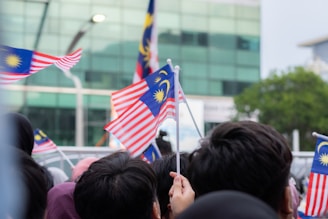 A crowd of people holding and waving Malaysian flags, which are predominantly red, white, blue, and yellow. The background consists of a glass building and green foliage.