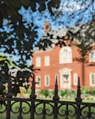 A wrought iron fence with pointed tips is prominently in the foreground, partially obscuring the view of a large red brick house with white window frames and a decorative front entrance. The background shows a garden with colorful flowers, and trees provide shade and framing.