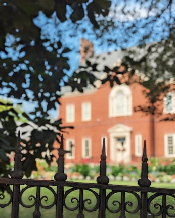 A wrought iron fence with pointed tips is prominently in the foreground, partially obscuring the view of a large red brick house with white window frames and a decorative front entrance. The background shows a garden with colorful flowers, and trees provide shade and framing.