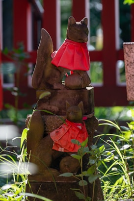 Two fox statues are adorned with bright red bibs, positioned outdoors with lush greenery surrounding them. The foxes are of different sizes, with one seated above the other. Sunlight filters through the leaves, casting dappled shadows on the statues.