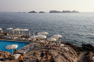 A seaside resort area with lounge chairs and umbrellas overlooking a clear blue swimming pool. People are sunbathing and enjoying the coastal view. The ocean is calm, with small, rocky islands visible in the distance under a hazy sky.