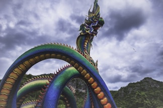 A vibrant, multi-colored serpent-like dragon statue with intricate scales is dramatically posed against a backdrop of dark, stormy clouds. The dragon features a fierce expression with sharp teeth, towering over lush greenery and mountainous landscape, evoking a sense of myth and grandeur.