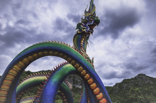 A vibrant, multi-colored serpent-like dragon statue with intricate scales is dramatically posed against a backdrop of dark, stormy clouds. The dragon features a fierce expression with sharp teeth, towering over lush greenery and mountainous landscape, evoking a sense of myth and grandeur.