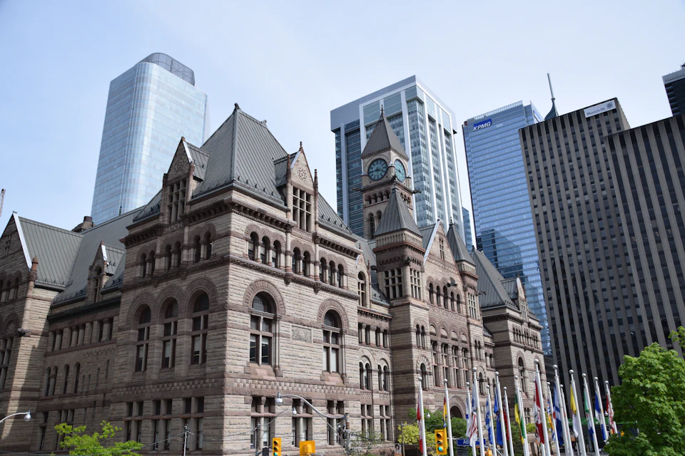 The image depicts a large, ornate stone building with a clock tower and multiple arched windows, situated on a street corner surrounded by modern skyscrapers. The building features a gray roof and is adorned with numerous flags fluttering in front of it. In the foreground, there are trees providing a natural element to the urban landscape.