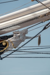 A ship's figurehead depicts a woman with flowing hair in a dynamic pose, reaching forward with a determined expression. Surrounding the figurehead are multiple ropes and chains, with a backdrop of blue sky. The figurehead has gold accents and is intricately carved.